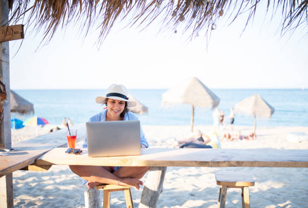 Shot of a happy tourist woman sitting at the beach cafe and using a laptop.